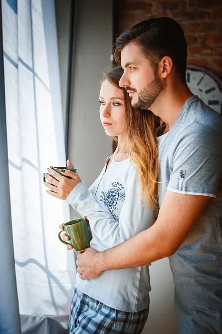 The couple by the window with mugs, he hugs her from behind