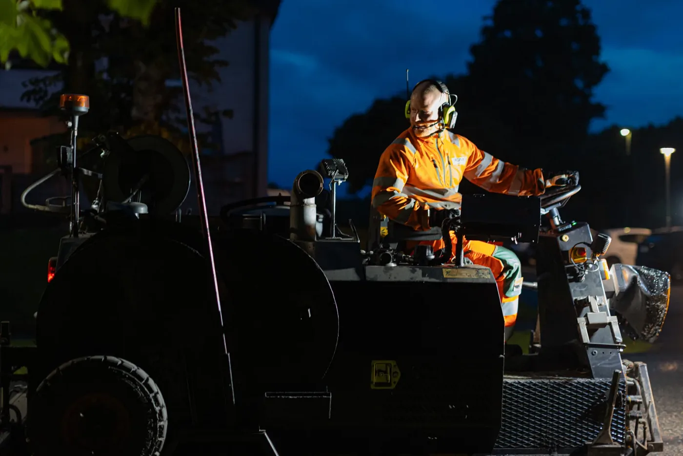 Maskinförare i varselkläder och hörselskydd vid spakarna på en asfaltmaskin på kvällen