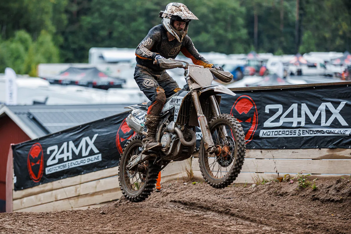 Muddy motocross rider jumping in front of a 24MX sponsor banner, Kinds